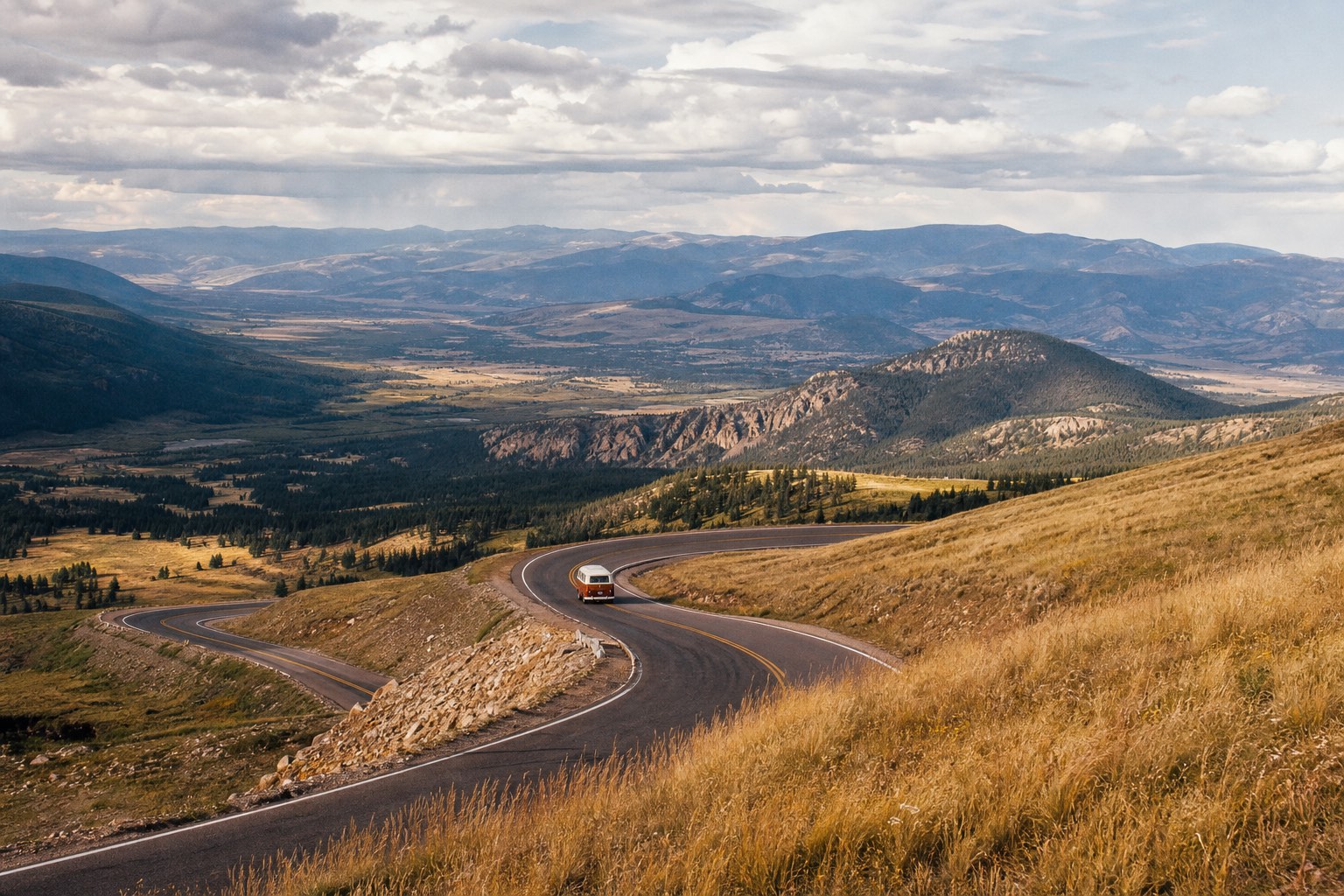 A rust-and-cream camper van descends from a mountain pass into a broad Western valley